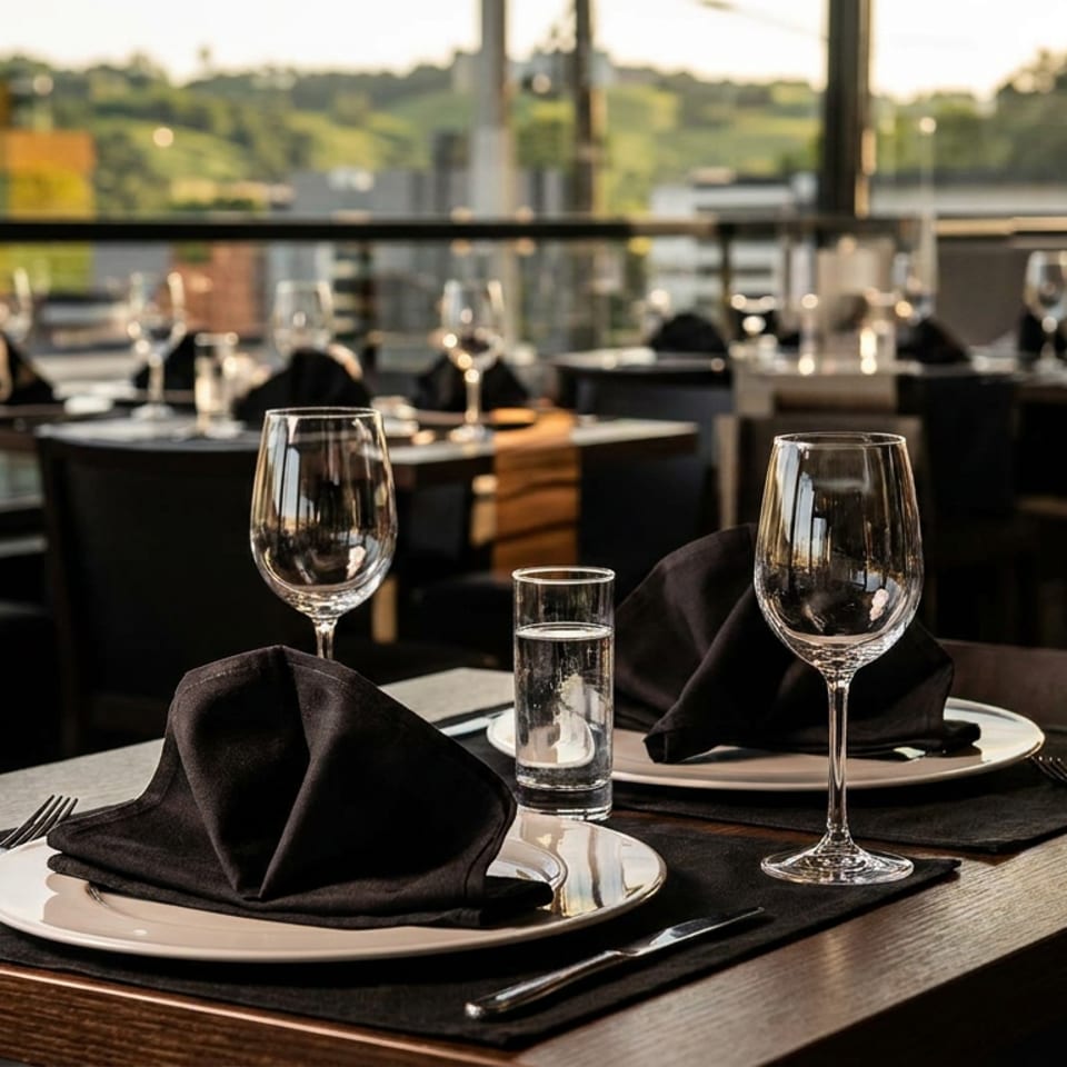 Elegant restaurant dining room with set tables, black folded napkins, wine glasses, water glasses, and large windows overlooking greenery.