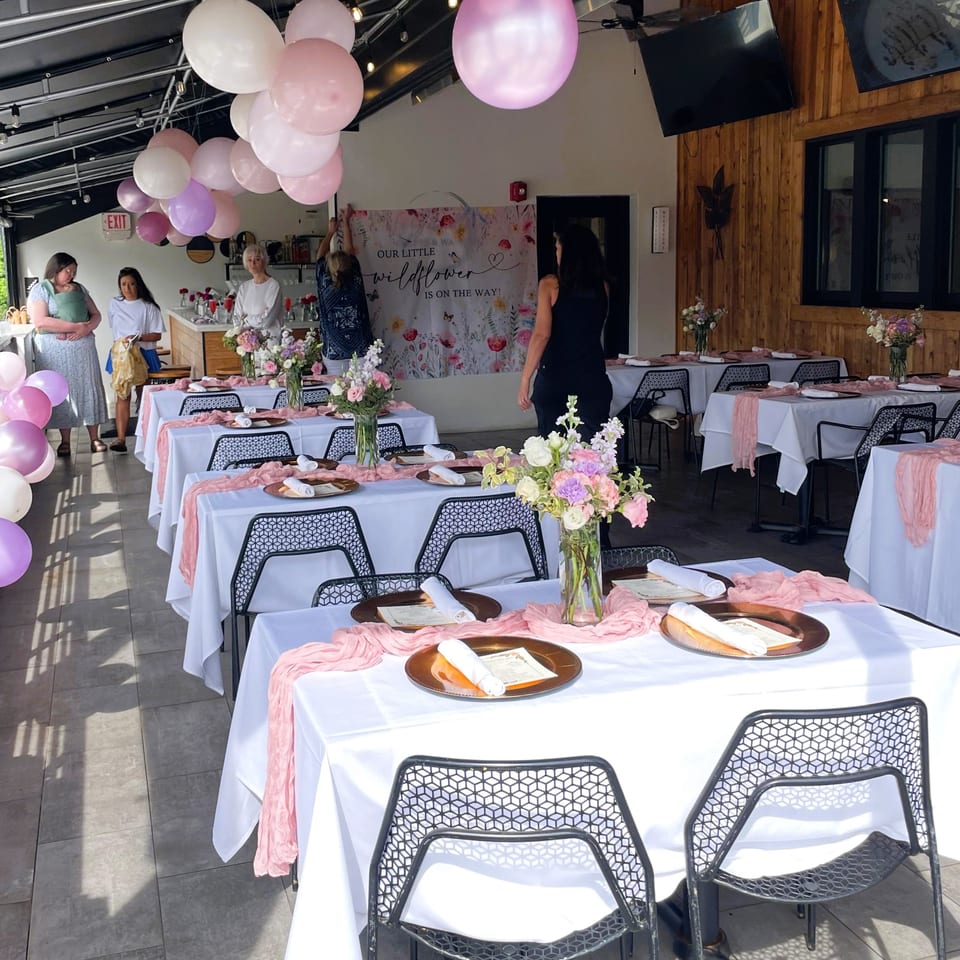 Restaurant dining room decorated with pink balloons, floral centerpieces, and white tables set for a wildflower-themed celebration.