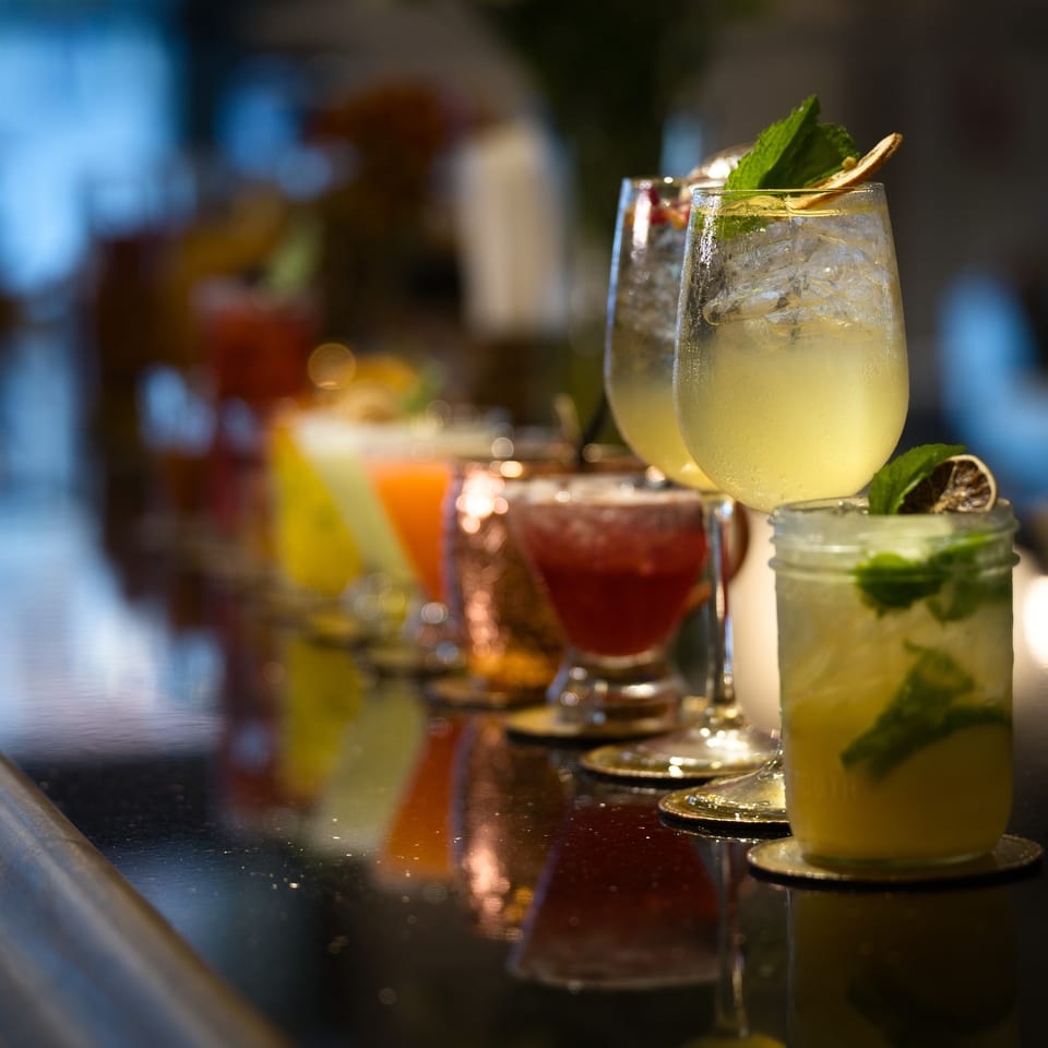 Colorful cocktails garnished with mint and citrus sit on a reflective bar counter in a dimly lit restaurant.