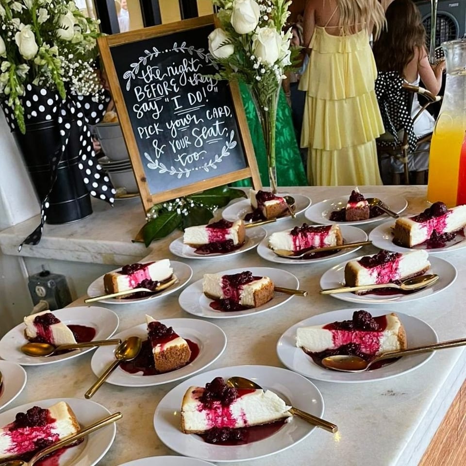 Plates of berry-topped cheesecake sit on a dessert table beside flowers, colorful drinks, and a wedding-themed chalkboard.