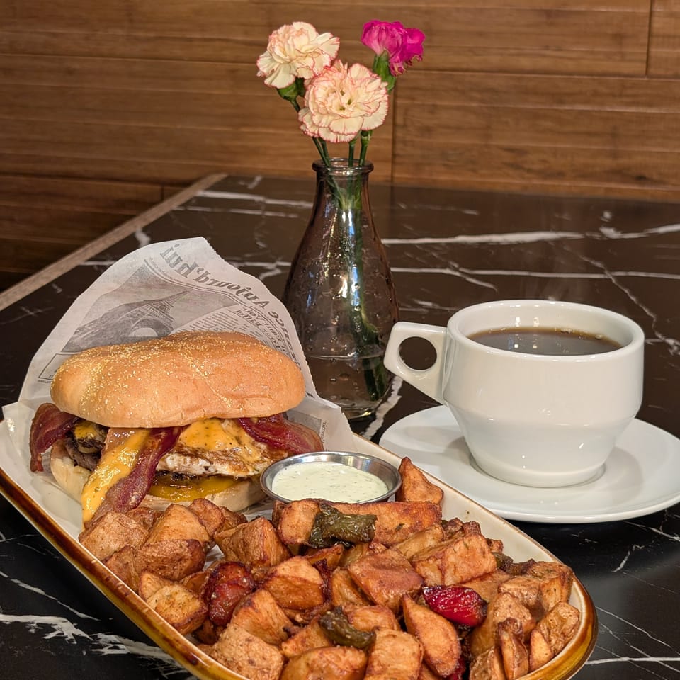 Bacon cheeseburger with roasted potatoes, dipping sauce, and coffee on a marble table beside a vase of carnations.