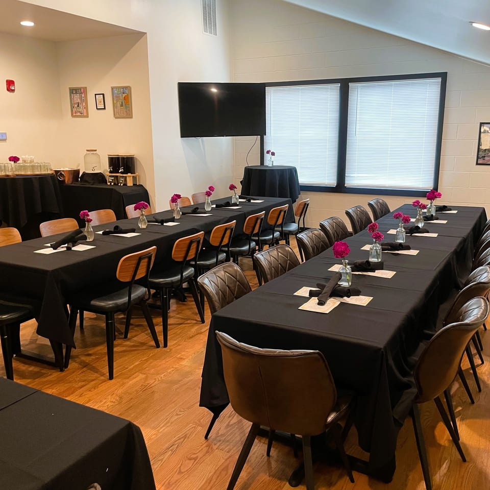 Restaurant dining room with black tablecloths, pink flower centerpieces, set place settings, buffet station, and wall-mounted television.