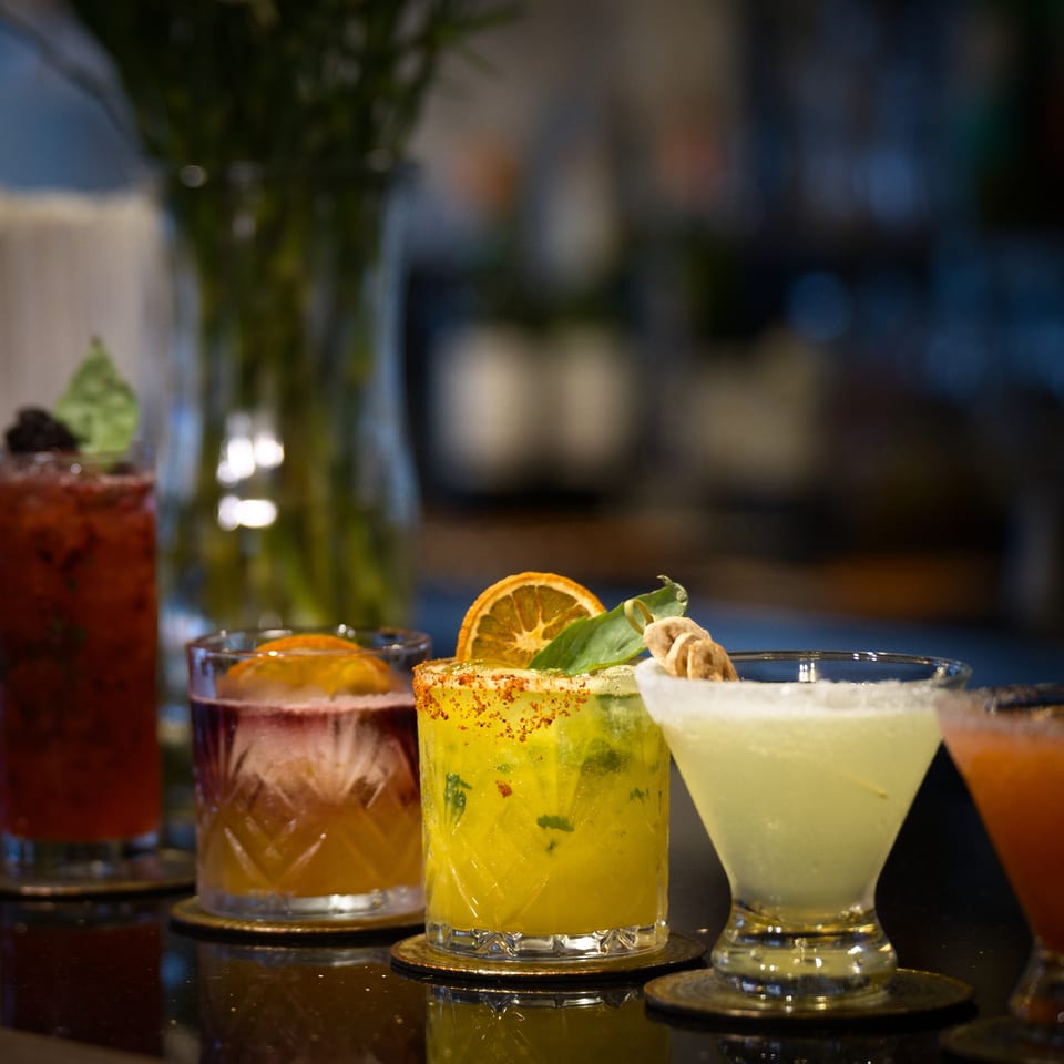 Colorful cocktails, including a chili-rimmed citrus drink, sit on coasters at a dimly lit restaurant bar.