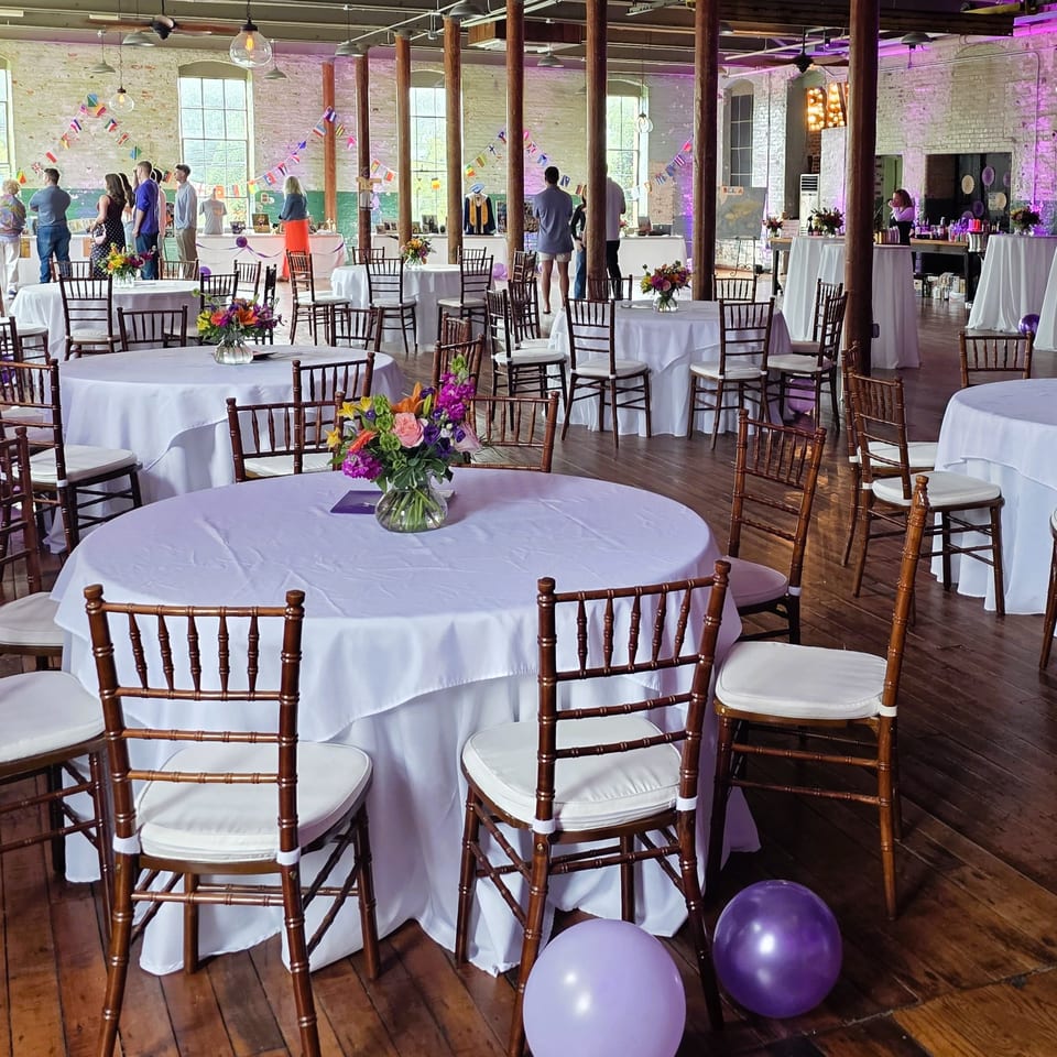 Spacious industrial event venue with white-clothed tables, floral centerpieces, wooden chairs, international flag bunting, and guests.