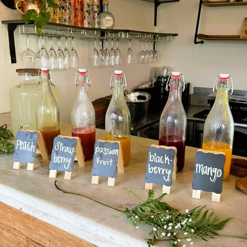 Five glass bottles of colorful fruit beverages labeled peach, strawberry, passion fruit, blackberry, and mango on a café counter.